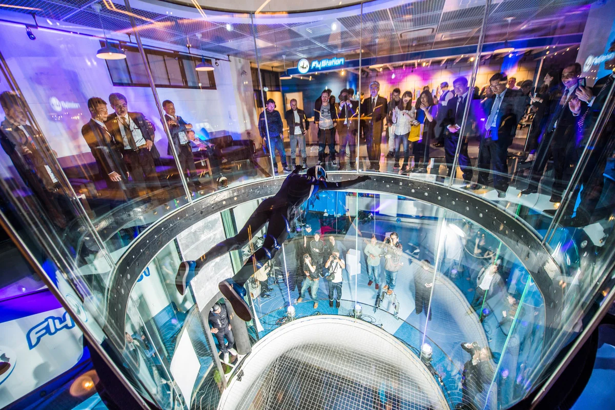 Profesyonel uçucu Leo Volkov, FlyStation Japan'daki TT45 PRO uçuş odası içinde performans sergiliyor. Yukarıdan aşağıya bakış açısı, silindirik çerçevesiz cam duvarları ve hava akışı sütununun tabanındaki paslanmaz çelik güvenlik ağını ortaya çıkarıyor. Seyirciler, odanın üst bölümünü çevreleyen yapısal cam zeminden uçuşu izliyor. 4,5 metre çapındaki bu rüzgâr tüneli, yüksek hızlı manevralar için görüş ve güvenlik sağlayan TunnelTech'in imzası niteliğindeki yuvarlak çok katmanlı cam tasarımına sahiptir.