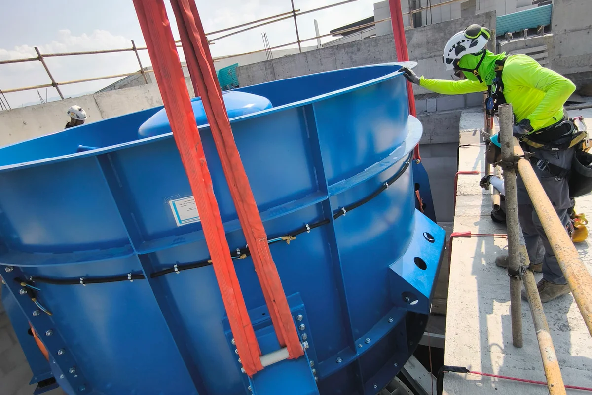 Bouwvakkers begeleiden de installatie van een krachtige axiale ventilatorunit voor een TunnelTech TT43 Smart windtunnel bij het China Flight Town project. De blauwe stalen ventilatorbehuizing, opgehangen aan zware kraanbanden, daalt af in de verticale betonnen schacht van het retourkanaal. Zichtbaar onder de behuizing is de waaier met koolstofvezelbladen, ontworpen voor efficiënte werking binnen het recirculerende luchtstroomcircuit.
