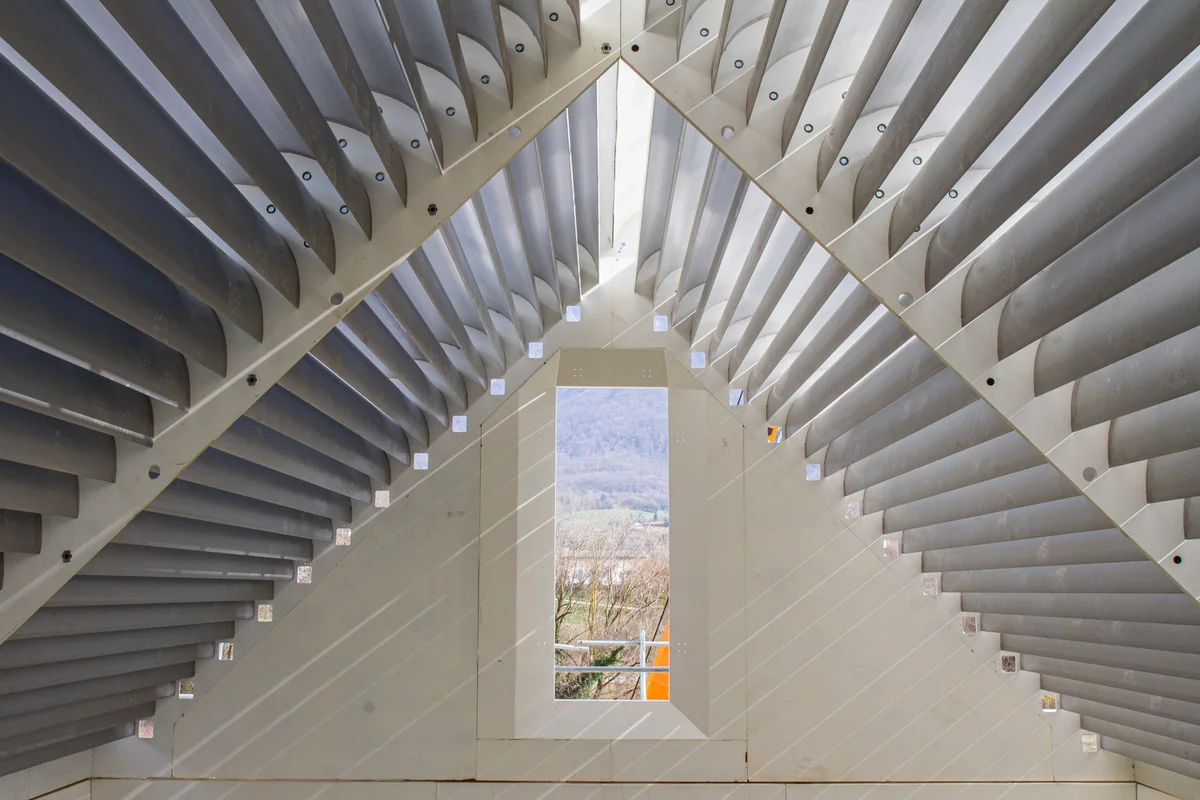 This image displays assembled aluminum turning vanes positioned at the top of the flight chamber in a double-loop wind tunnel at the Windalps facility. These vanes form a triangular, house-like structure designed to split the vertical airflow into two separate paths, directing the air into the left and right return ducts. The facility is shown prior to the installation of the roof, allowing sunlight to illuminate the vane structure and internal coolant channels.