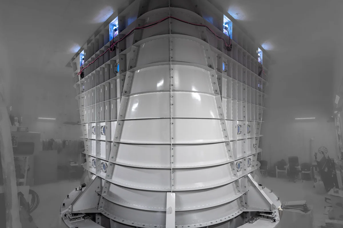 De afbeelding toont de installatie van de stalen diffuserassemblage voor een TunnelTech TT52 Pro windtunnel in de Kuzbas Arena-faciliteit. Dit uitdijende kanaalcomponent bevindt zich direct boven de vluchtkamer om de luchtstroom te vertragen en druk te herstellen binnen het recirculerende circuit. De structuur bestaat uit gesegmenteerde witte stalen panelen met externe structurele ribben en geboute flensverbindingen, die de bovenste overgangssectie vormen van het verticale luchtstroomcircuit met een diameter van 5,2 meter.