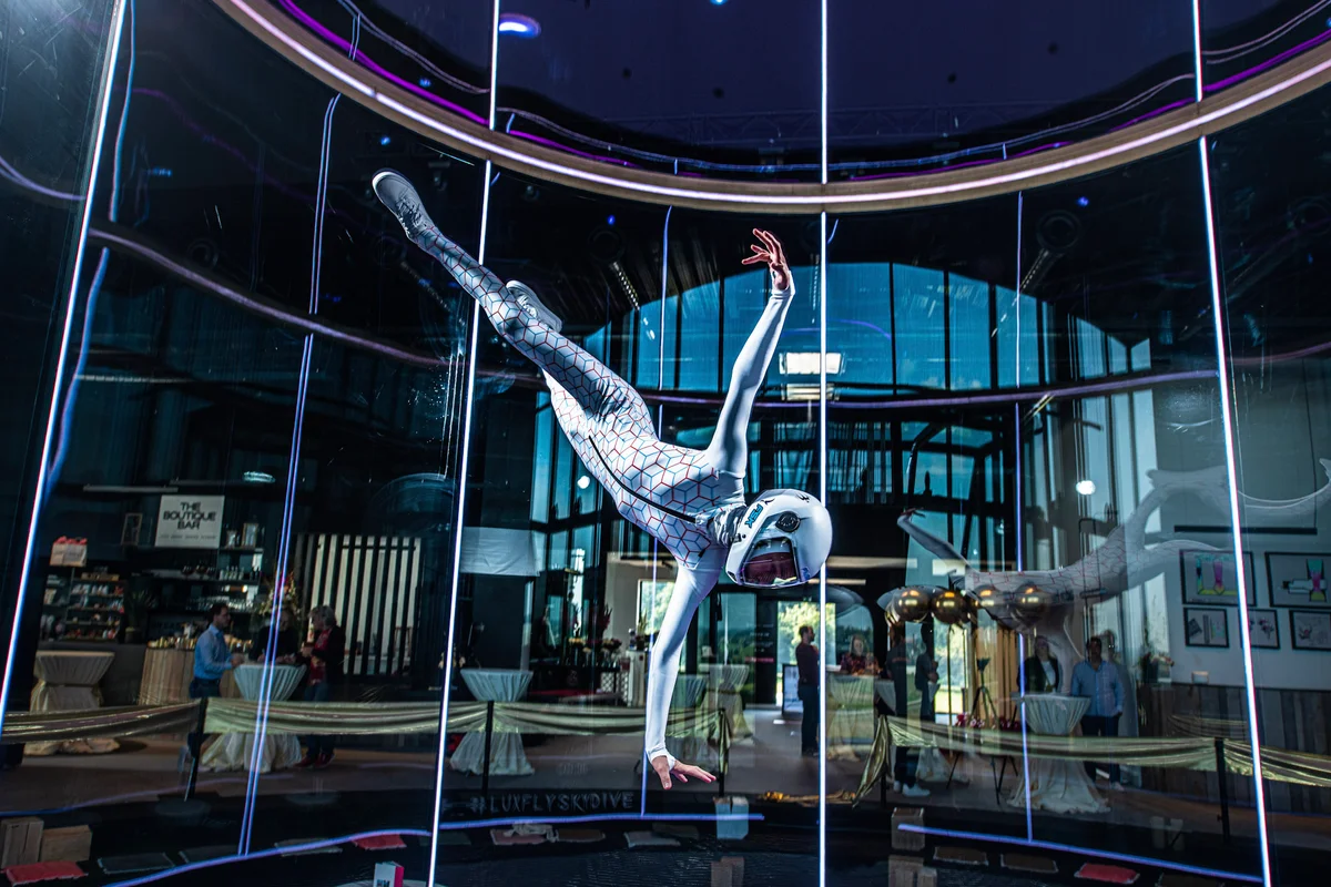 Feith Máté führt ein dynamisches Manöver in der Flugkammer der Luxfly Indoor-Skydiving-Anlage in Luxemburg aus. Diese Anlage nutzt einen TunnelTech TT45 PRO Windkanal mit einer Glas-Flugkammer von 4,5 Metern Durchmesser. Die transparenten, mehrschichtigen und schallabsorbierenden Glaswände ermöglichen es den Zuschauern im angrenzenden Barbereich, den Flug direkt zu beobachten. Vertikale LED-Lichtstreifen beleuchten die Begrenzungen der Kammer, während das Sicherheitsnetz aus Stahlseilen an der Basis der Luftstromsäule sichtbar ist.