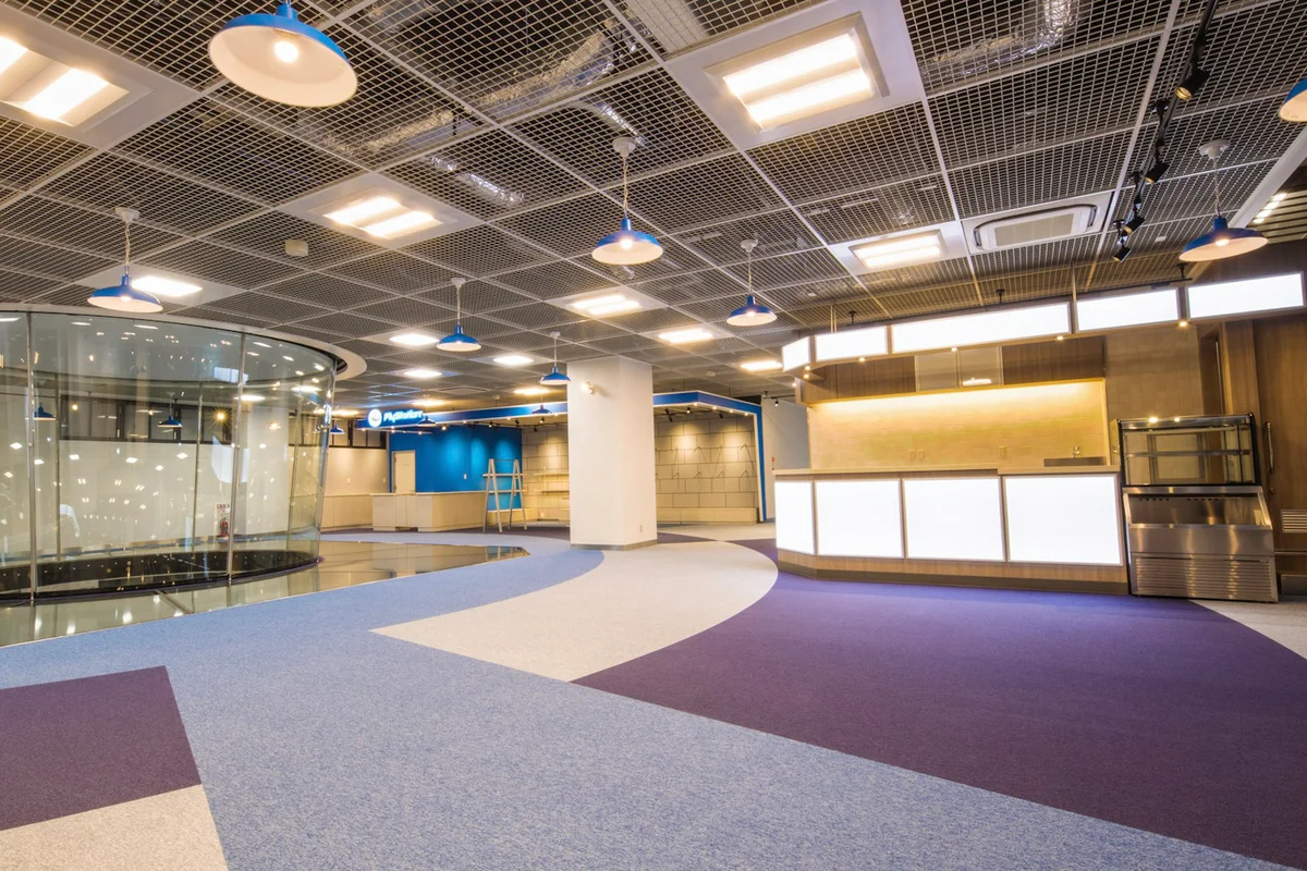 Het interieur van de tweede verdieping van de FlyStation Japan-faciliteit toont het bovenste gedeelte van de TT45 Pro vluchtkamer. Een structurele glazen vloer omringt de bovenkant van de kamer. Aan de rechterkant bevindt zich een cafégedeelte met een toonbank met verlichte witte panelen. De achtergrond toont de FlyStation-branding en schappen voor een merchandise-winkel. Het plafond bestaat uit een open rooster met hanglampen, uitkijkend op een vloer met geometrisch blauw en paars tapijt. De receptie bevindt zich op de eerste verdieping.