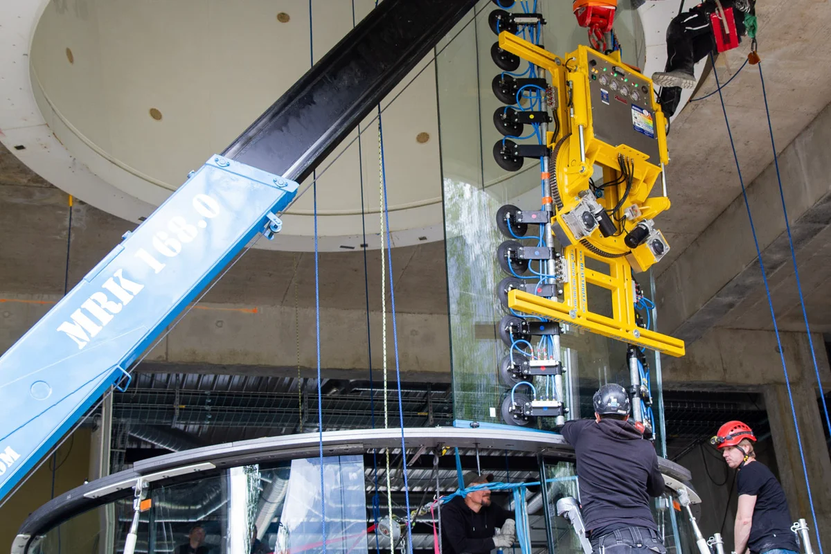 En spesialisert edderkoppkran utstyrt med en kraftig vakuumløfter posisjonerer et stort buet glasspanel for TT45 PRO flykammer. Industriklatrere som henger fra den øvre betongringen leder det flerlags støyabsorberende glasset inn i stålrammen, mens teknikere på stillaser justerer basen. Denne monteringsprosessen ved Wind Alps-anlegget i Frankrike konstruerer det gjennomsiktige, sylindriske flyområdet til vindtunneln med lukket krets.