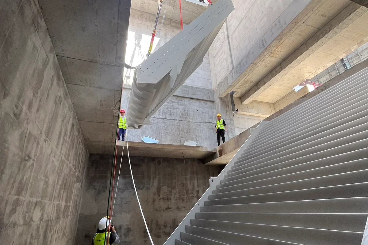 Teknisi memandu pemasangan sirip pengarah dengan pendinginan terintegrasi ke dalam struktur saluran beton terowongan angin TT43 Smart di China Flight Town. Sebuah derek menurunkan segmen sirip aluminium melalui bukaan atas ke dalam saluran balik. Sirip pengarah ini menggabungkan saluran pendingin internal untuk berfungsi sebagai penukar panas, mengelola suhu udara sambil mengarahkan kembali aliran udara 90 derajat dengan turbulensi dan kehilangan tekanan minimal.