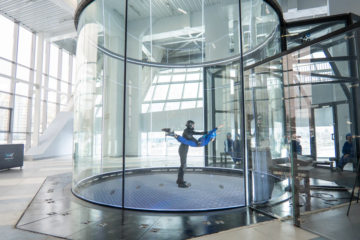 Isang instructor ang nag-i-spot sa flyer habang naka-belly flying sa loob ng TT52 Pro wind tunnel sa Kuzbass Arena. Ang 5.2-meter flight chamber na ito ay may frameless, noise-absorbing glass para sa perfect flow uniformity at malinaw na visibility. Makikita ang low-drag steel cable safety net sa ilalim, at ang glass airlock system sa kanan para sa pagpasok.