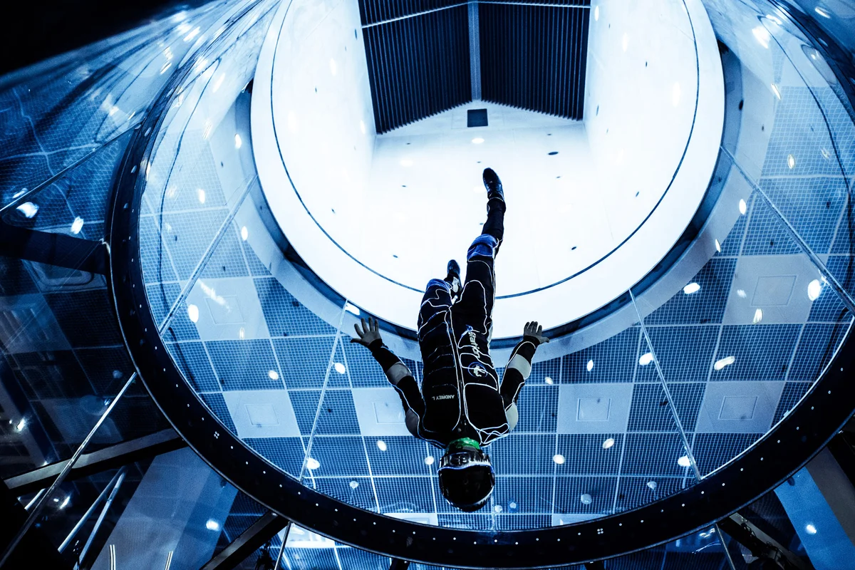 Ein professioneller Flyer führt ein Head-Down-Manöver in der gläsernen Flugkammer des TT45 PRO Windkanals bei FlyStation Japan aus. Die Perspektive nach oben zeigt den Übergang von der transparenten Flugstrecke zum Diffusor und der oberen Kanalecke, die mit parallelen Umlenkleitblechen ausgestattet ist. Der Flyer trägt einen Markenanzug, während er sich im schnellen vertikalen Luftstrom bewegt. Die Kammer mit 4,5 Metern Durchmesser verfügt über gebogene Mehrschichtglas-Paneele, die für Sichtbarkeit und Sicherheit bei fortgeschrittenen Flugoperationen ausgelegt sind.