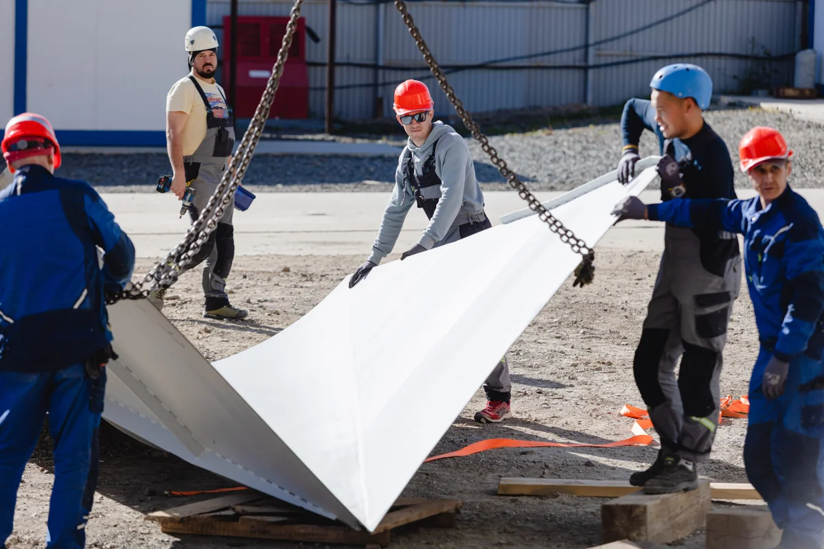 Kru instalasi khusus TunnelTech memanuver panel difuser putih besar yang digantung dengan rantai derek. Tim mengarahkan bagian duct komposit FRP bersudut tersebut ke posisinya untuk perakitan di lokasi konstruksi. Komponen struktural ini membentuk bagian penting dari sirkuit aerodinamis terowongan angin, yang dirancang khusus untuk memperluas aliran udara dan memulihkan tekanan di hilir ruang terbang. Para pekerja mengenakan alat pelindung diri standar saat menangani beban berat tersebut.