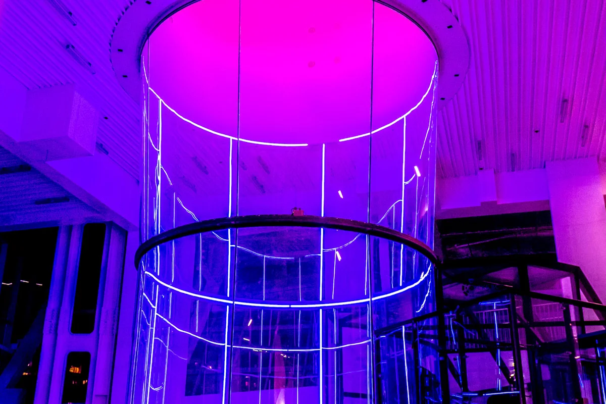 Ein beeindruckender Blick auf den TT52 Pro in der Kuzbass Arena mit seiner massiven 5,2-Meter-Flugkammer. Der zylindrische transparente Abschnitt aus mehrschichtigem Schallschutzglas wird stimmungsvoll violett beleuchtet und mündet nahtlos in den oberen Rückführkanal. Rechts im Bild sind die verglaste Vorkammer und das Luftschleusensystem zu erkennen. Die Anlage nutzt ein effizientes Single-Loop-Umlaufdesign, das speziell für hohen Durchsatz und professionelles Flugtraining entwickelt wurde.