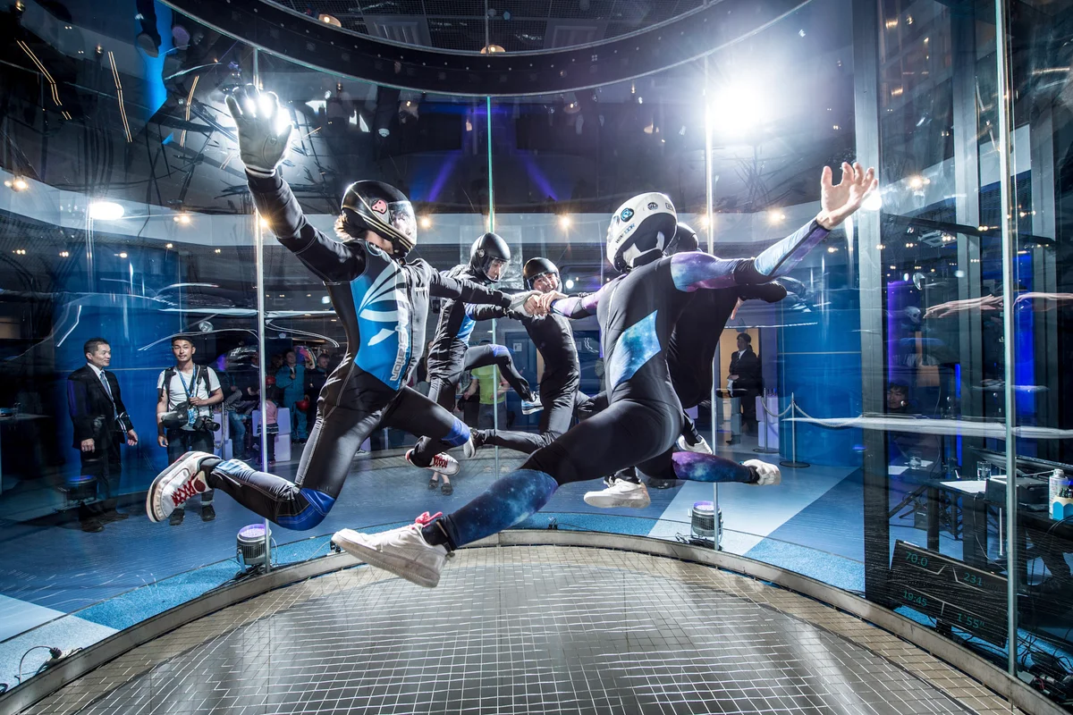Four professional flyers execute a coordinated head-up formation inside the TT45 PRO wind tunnel at FlyStation Japan. The group maintains a sit-fly orientation above the tensioned steel cable safety net. The flight chamber features TunnelTech's signature frameless conical glass section, offering unobstructed visibility for spectators observing from the surrounding area. A digital display panel mounted inside the chamber provides real-time telemetry data during the performance.