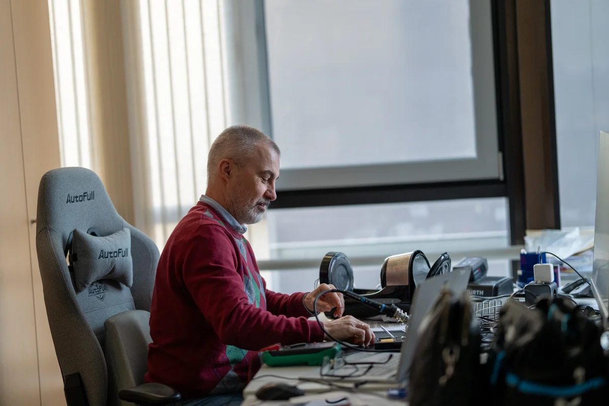 A TunnelTech mérnöke egy elektronikai összeszerelésre és tesztelésre felszerelt asztalnál dolgozik. A munkaterületen forrasztópáka, digitális multiméter és a hardver karbantartására vagy fejlesztésére szolgáló precíziós szerszámok találhatók. A mérnök az áramköri alkatrészekre összpontosít, támogatva a vállalat belső gyártását a szélcsatorna-műveletekhez szükséges vezérlőelektronika és automatizálási rendszerek terén.