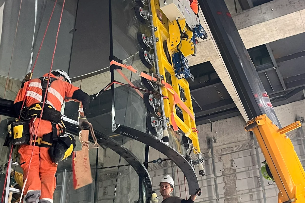 Des techniciens installent un panneau de verre incurvé pour la chambre de vol d'une soufflerie TT43 Smart sur le projet China Flight Town. Un palonnier à ventouses haute résistance fixé à une grue positionne la section en verre multicouche insonorisant dans la structure en acier. L'équipe d'installation utilise des techniques d'accès sur cordes et des échelles pour aligner le verre chimiquement trempé, assurant un ajustement précis pour la zone de vol de 4.3 mètres de diamètre.