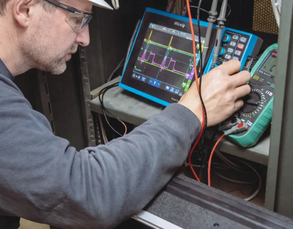 A service technician performs electrical diagnostics on the wind tunnel's industrial automation and control systems. The engineer uses a Micsig tablet oscilloscope to measure signal waveforms and verify the performance of the electronic components. The open equipment rack contains DIN-rail mounted interface modules and wiring terminals. This maintenance activity supports the reliability of the internal control electronics and software developed by TunnelTech's engineering team.
