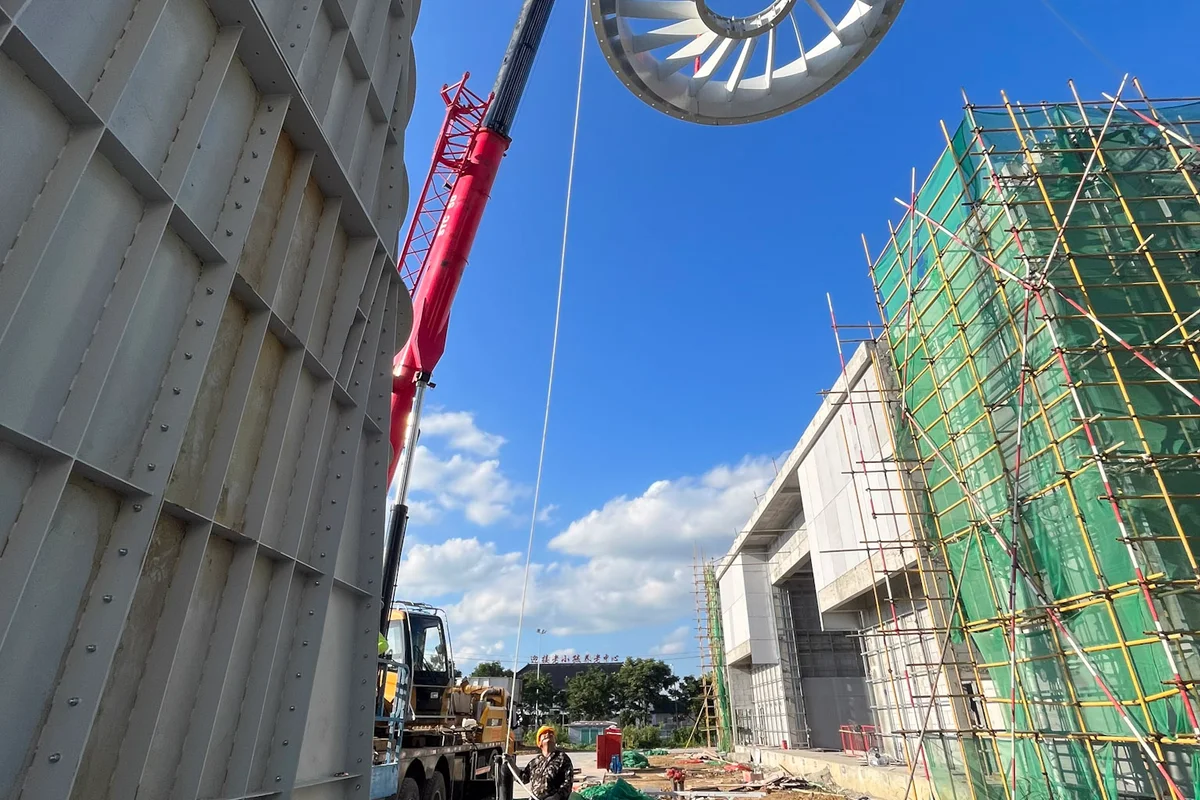 Ein Schwerlastkran hebt einen kreisförmigen Stator-Leitschaufelring während der Installation eines TT43 Smart Windkanals beim Flight Town-Projekt in China in Position. Diese Komponente ist Teil der Axialventilator-Baugruppe und verfügt über feststehende Leitschaufeln, die dazu dienen, den Luftstrom im Kreislauf gleichzurichten. Links steht ein großer vorgefertigter stählerner Luftkanalabschnitt zur Integration in die Windkanalstruktur bereit.