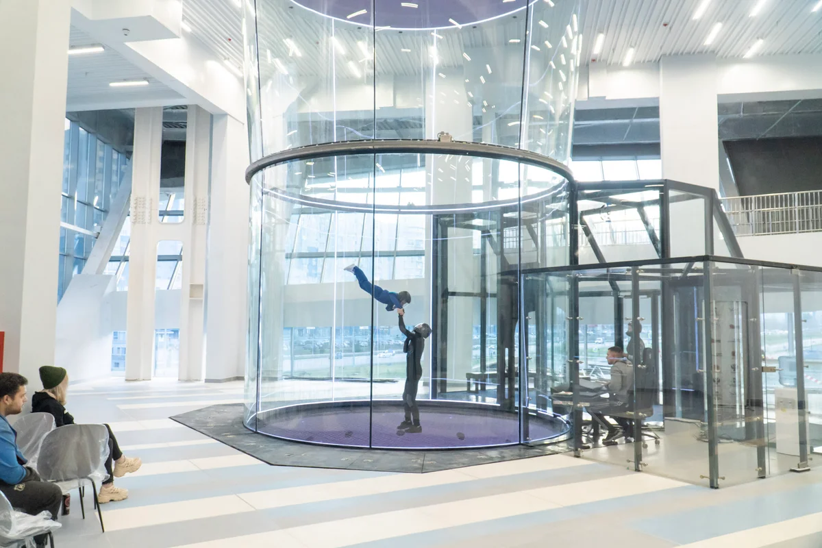 Démonstration de sécurité dans la chambre de vol (flight chamber) TT52 Pro de la Kuzbas Arena : un instructeur assiste un enfant en position de belly flying. Ce tunnel de 5,2 mètres de diamètre se distingue par ses parois en verre multicouche (multilayer glass) sans cadre, offrant une transparence totale pour les spectateurs de l'atrium. Sur la droite, le poste de l'opérateur jouxte le sas (airlock) vitré, assurant une gestion précise du flux d'air (airflow).