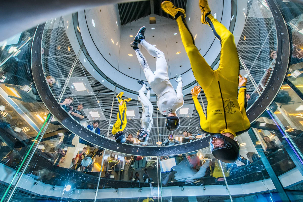 Four athletes execute head-down maneuvers inside the FlyStation Japan wind tunnel during the Sakura Cup. This TT45 Pro installation features a 4.5-meter diameter flight chamber with perfectly round multilayer glass walls. The image is taken from below the transparent glass floor, looking upward past the flyers toward the upper diffuser and return air intake. Spectators observe the dynamic flying routine from the surrounding viewing deck.