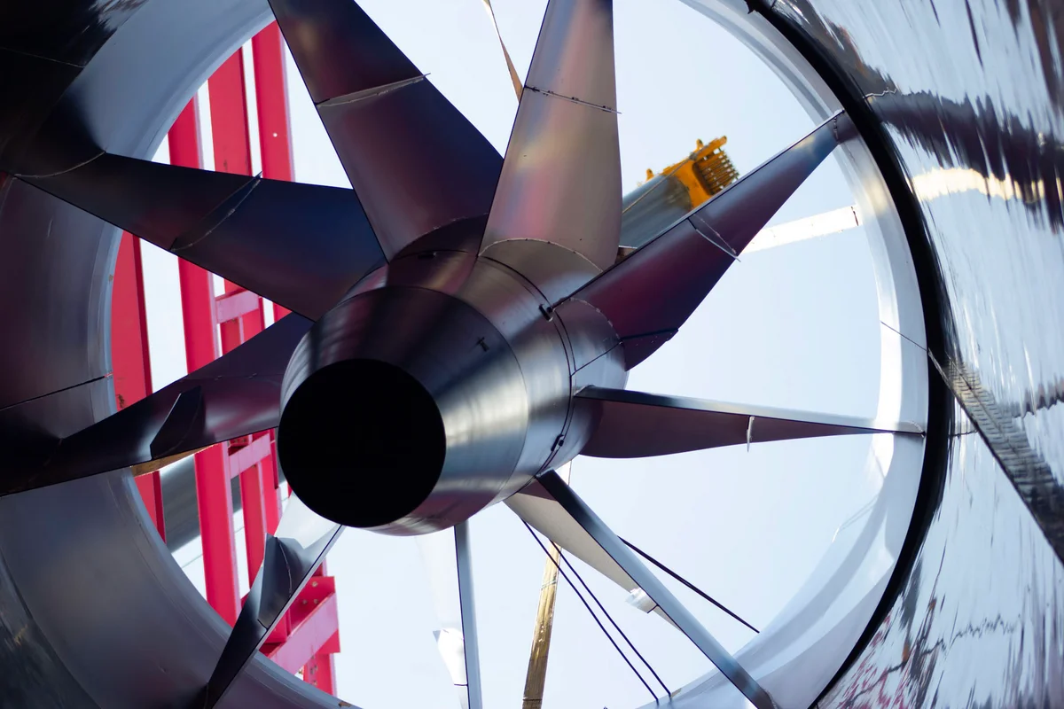 Billedet fanger installationen af en hoved-aksialventilator til Luxfly-vindtunnelen i Luxembourg. Enheden har en rotor med stor diameter og kulfiberblade monteret på en vertikal akse. En kran placerer ventilatorkomponenten i luftkanalen, omgivet af anlæggets røde stålstøttestruktur. Denne komponent driver luftflowet for et TunnelTech TT45 PRO-system.