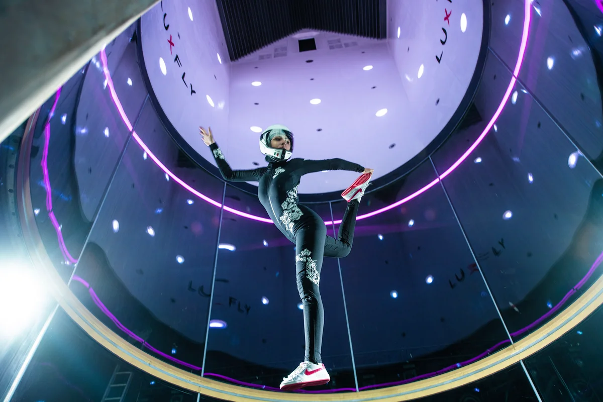 Flyerka provádí freestyle manévr uvnitř letové komory zařízení pro indoor skydiving Luxfly v Lucembursku. Zařízení využívá větrný tunel TunnelTech TT45 Pro s vysokou, bezrámovou válcovou skleněnou částí navrženou pro maximální viditelnost a bezpečnost. Dokonale kulaté stěny z vícevrstvého skla obklopují letový prostor, kde atletka udržuje vertikální orientaci. Fialové LED osvětlení obklopuje horní oblast vedoucí k difuzoru a zvýrazňuje značku LUXFLY na vnitřních panelech.