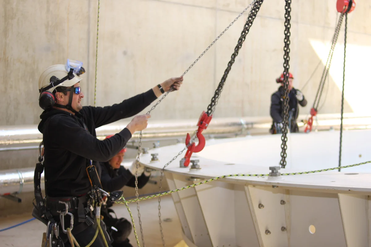 I tecnici di installazione TunnelTech eseguono operazioni di rigging durante l'assemblaggio di una galleria del vento TT45 PRO. I membri del team, indossando imbracature di sicurezza complete e dispositivi di protezione, utilizzano paranchi a catena per posizionare una grande sezione curva di condotto in acciaio all'interno della struttura in calcestruzzo dell'impianto. Questa fase di costruzione prevede l'allineamento preciso e l'imbullonatura di pesanti flange strutturali per formare il circuito aerodinamico continuo richiesto per il sistema della camera di volo da 4.5 metri.