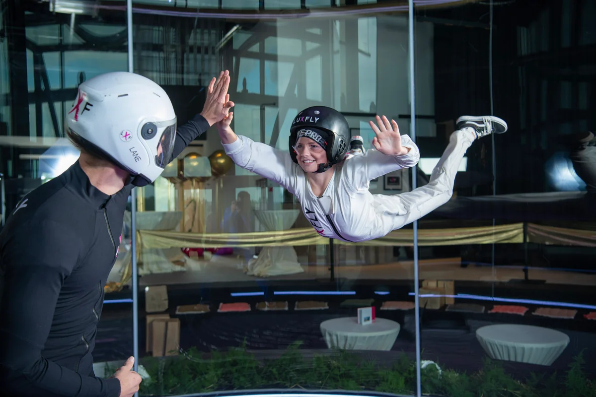 Ein Instruktor und ein Flyer in Belly-Down-Ausrichtung interagieren in der Flugkammer der Indoor-Skydiving-Anlage Luxfly in Luxemburg. Diese Anlage verfügt über das Windkanalmodell TunnelTech TT45 PRO. Das Bild zeigt die perfekt runden Wände aus Mehrschichtglas, die den Zuschauern eine klare Sicht bieten. Der Instruktor steht auf dem Stahlseil-Sicherheitsnetz, während der Flyer eine stabile neutrale Körperposition im vertikalen Luftstrom beibehält.