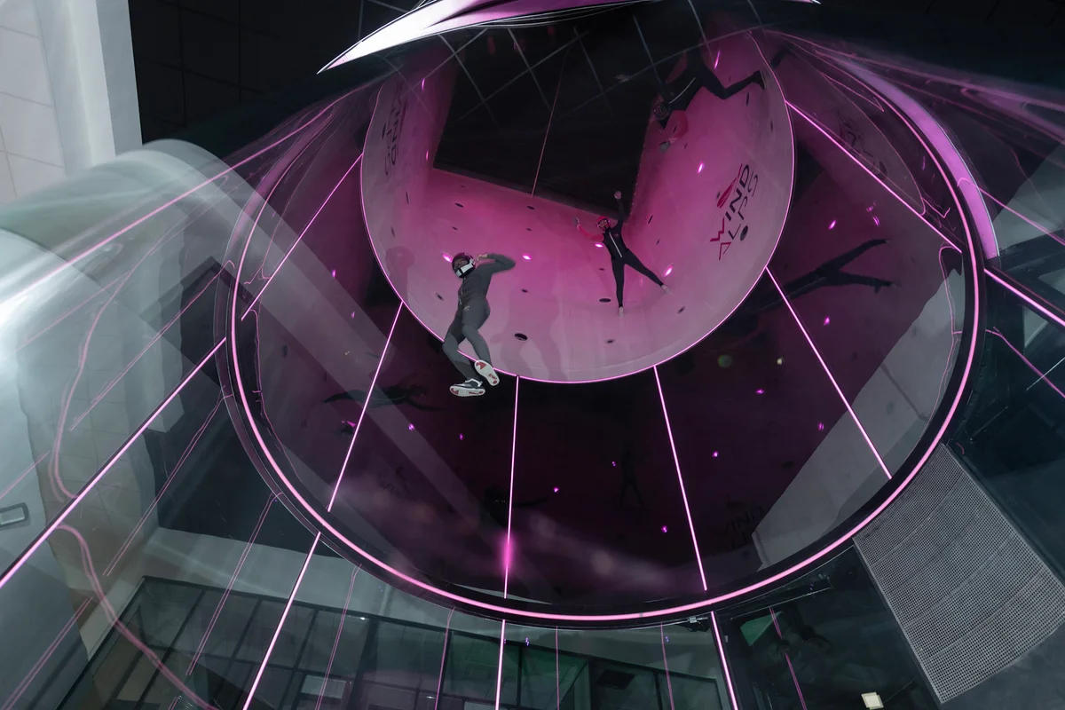 A vertical perspective inside the TT45 PRO flight chamber at Wind Alps, France. Three flyers are practicing head-up maneuvers within the perfectly round, multilayer glass walls. Illuminated in pink, this 4.5-meter ProFly installation demonstrates the clarity and safety of TunnelTech's frameless glass technology, designed for high-speed advanced freefly disciplines.