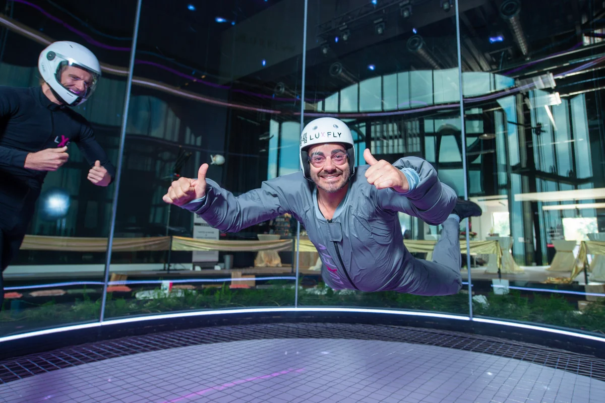 Ein Teilnehmer in einem grauen Fluganzug führt ein Belly Flying Manöver in der Luxfly Indoor-Skydiving Anlage aus. Ein Instruktor überwacht die Session im Hintergrund innerhalb der TT45 PRO Flugkammer. Das Bild zeigt den Boden aus gespanntem Stahlseil-Sicherheitsnetz und die perfekt runden Wände aus Mehrschichtglas, die den Flugbereich umschließen. Diese Kammer mit 4.3 Metern Durchmesser nutzt die rahmenlose Glastechnologie von TunnelTech, um eine klare Sicht auf den umgebenden Raum zu bieten und gleichzeitig einen konstanten Luftvolumenstrom aufrechtzuerhalten.