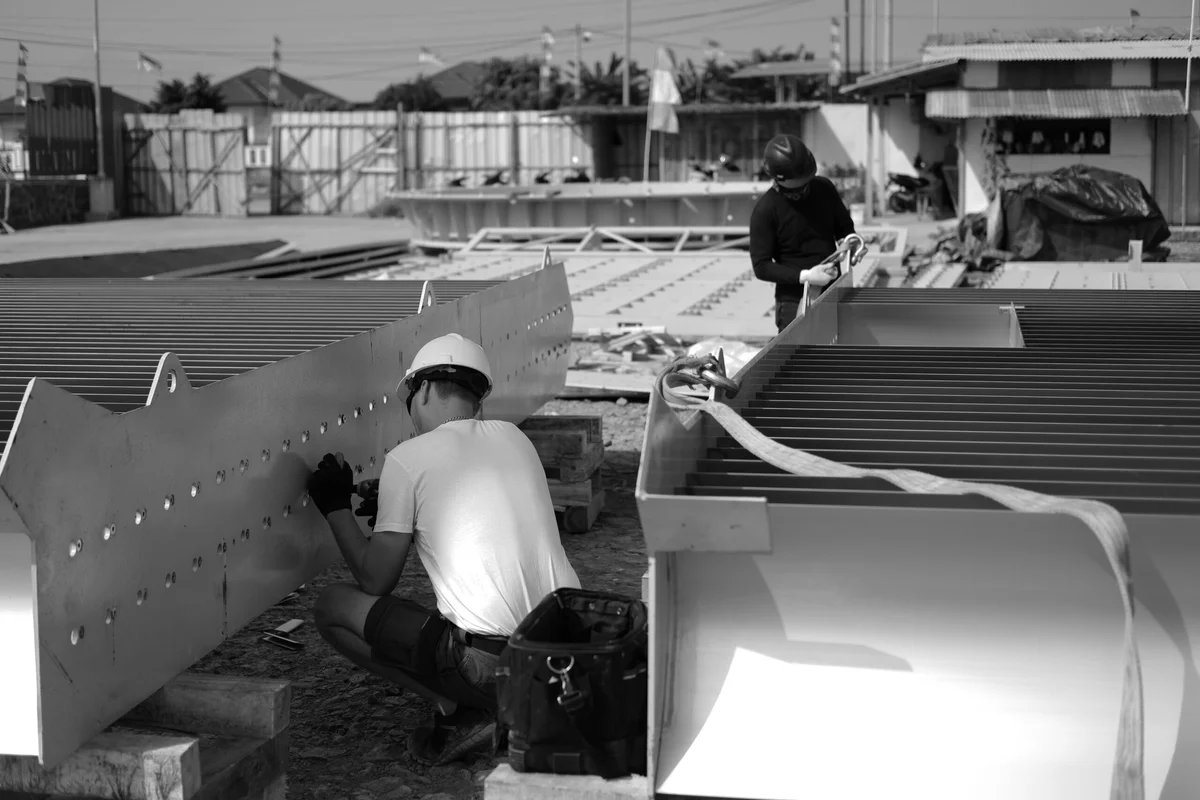 טכנאי ההתקנה של TunnelTech מרכיבים מקטעי תעלות פינתיים גדולים המכילים להבי הנחיה באתר בנייה חיצוני. הצוות מאבטח את מעטפת הפלדה ואת מבני הלהבים הפנימיים, הכוללים תעלות קירור אקטיבי משולבות לניהול תרמי. להבי הנחיה אלו מכוונים את ספיקת האוויר דרך פניות של 90 מעלות כדי להפחית הפסד לחץ וטורבולנציה תוך תפקוד כמחליפי חום. התמונה מציגה את הצוות מכין את המקטעים התעשייתיים הכבדים לשילוב בלולאת החזרה של מנהרת הרוח.