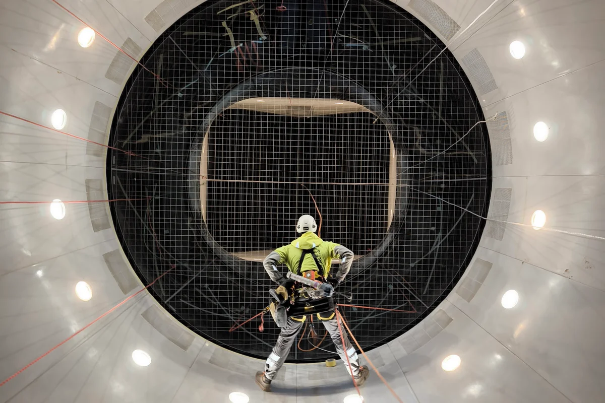 Ein Seilzugangstechniker hängt unter dem Sicherheitsnetz und führt Installationsarbeiten an der Kontraktionsstrecke durch. Die Perspektive blickt durch das Stahlkabelnetz und die Flugkammer nach oben und bietet einen Blick auf die Umlenkleitbleche am oberen Ende des Kreislaufs. Der Techniker befindet sich im weißen Komposit-Kontraktionskonus und nutzt eine Takelage, um die Fläche unterhalb des Flugdecks zu erreichen.