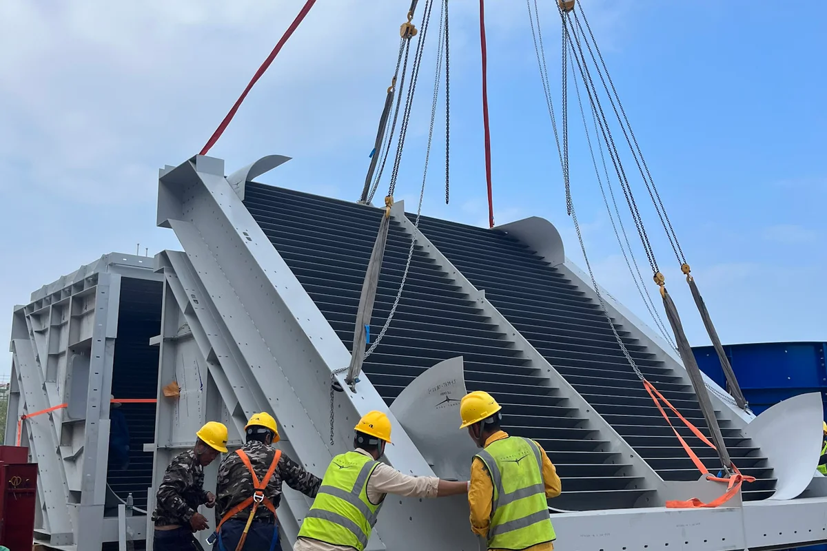 Uma grua levanta uma secção de aletas guia durante a instalação de um túnel de vento TT43 Smart no projeto China Flight Town. O pessoal da construção guia a unidade com estrutura de aço, que contém canais de refrigerante integrados para o sistema de arrefecimento ativo da instalação. Estas aletas funcionam como permutadores de calor para regular a temperatura do fluxo de ar enquanto redirecionam o ar através do canto do circuito de condutas de recirculação.