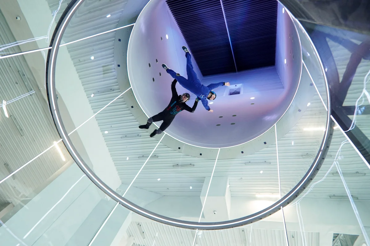 Ein Blick senkrecht nach oben durch die 5,2-Meter-Flugkammer zeigt einen Instruktor, der einen Anfänger auf einen hohen Taxi-Flug mitnimmt. Die perfekt runden Glaswände – ein Markenzeichen von TunnelTech – gehen nahtlos in den weißen Diffusor über, wobei die Umlenkleitbleche am Scheitelpunkt des Loops sichtbar sind. Integrierte Lichtringe sorgen für eine optimale Ausleuchtung während des Aufstiegs.