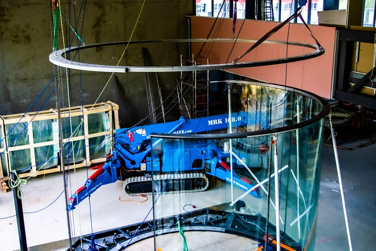 L'assemblage de la chambre de vol TT45 PRO a lieu sur le site de Wind Alps. Une grue araignée positionne les panneaux de verre multicouche incurvés sur la base en acier structurel pour former la zone de vol ronde et sans cadre. Des câbles de gréement suspendent l'anneau métallique supérieur tandis que des caisses supplémentaires contenant des sections de verre sont prêtes pour l'installation en arrière-plan. Cette phase de construction établit la zone de vol transparente caractéristique du modèle TT45 PRO.