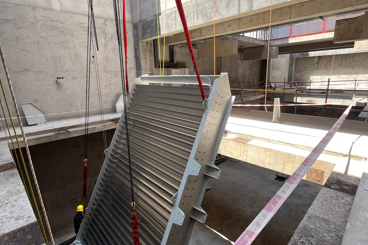 Installation of a turning vane assembly with integrated active cooling at the China Flight Town project. This component belongs to a TT43 Smart wind tunnel and combines airflow redirection with thermal management capabilities. The aluminum vanes contain internal coolant channels to dissipate heat generated during operation. The assembly is suspended by crane rigging within the concrete facility structure, ready for placement in the return duct corner.