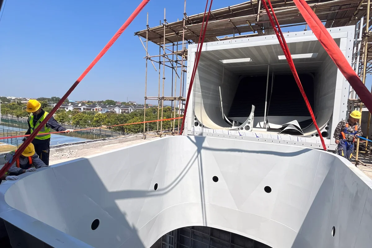 Installation of the TT43 Smart wind tunnel at China Flight Town proceeds with the lifting of a flight chamber diffuser section. The white component features a circular aperture that connects to the flight area. In the background, a horizontal return duct section equipped with black turning vanes rests on the structural steel scaffolding. Construction personnel manage the rigging straps to align the heavy aerodynamic segment.