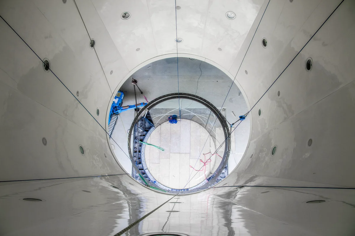 Perspektif vertikal melihat ke bawah melalui bagian difuser atas terowongan angin TT45 PRO di fasilitas Wind Alps. Dinding panel putih sirkuit aliran udara memiliki port akses melingkar dan mengarah turun ke tingkat flight chamber. Di bawah, rantai rigging dan mesin konstruksi biru membantu instalasi komponen ruang terbang kaca dan flensa koneksi baja.