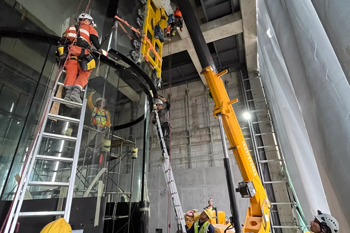 Technici TunnelTech instalují zakřivené panely z vícevrstvého skla pro letovou komoru větrného tunelu TT43 Smart v zařízení China Flight Town. Pavoučí jeřáb vybavený vakuovým zvedákem umisťuje těžký skleněný segment, zatímco tým využívá lanový přístup a žebříky pro přesné zarovnání. Instalační proces sestavuje válcovou letovou komoru o průměru 4,3 metru s využitím chemicky zpevněného skla pro zajištění strukturální bezpečnosti a konzistentní aerodynamiky průtoku vzduchu.