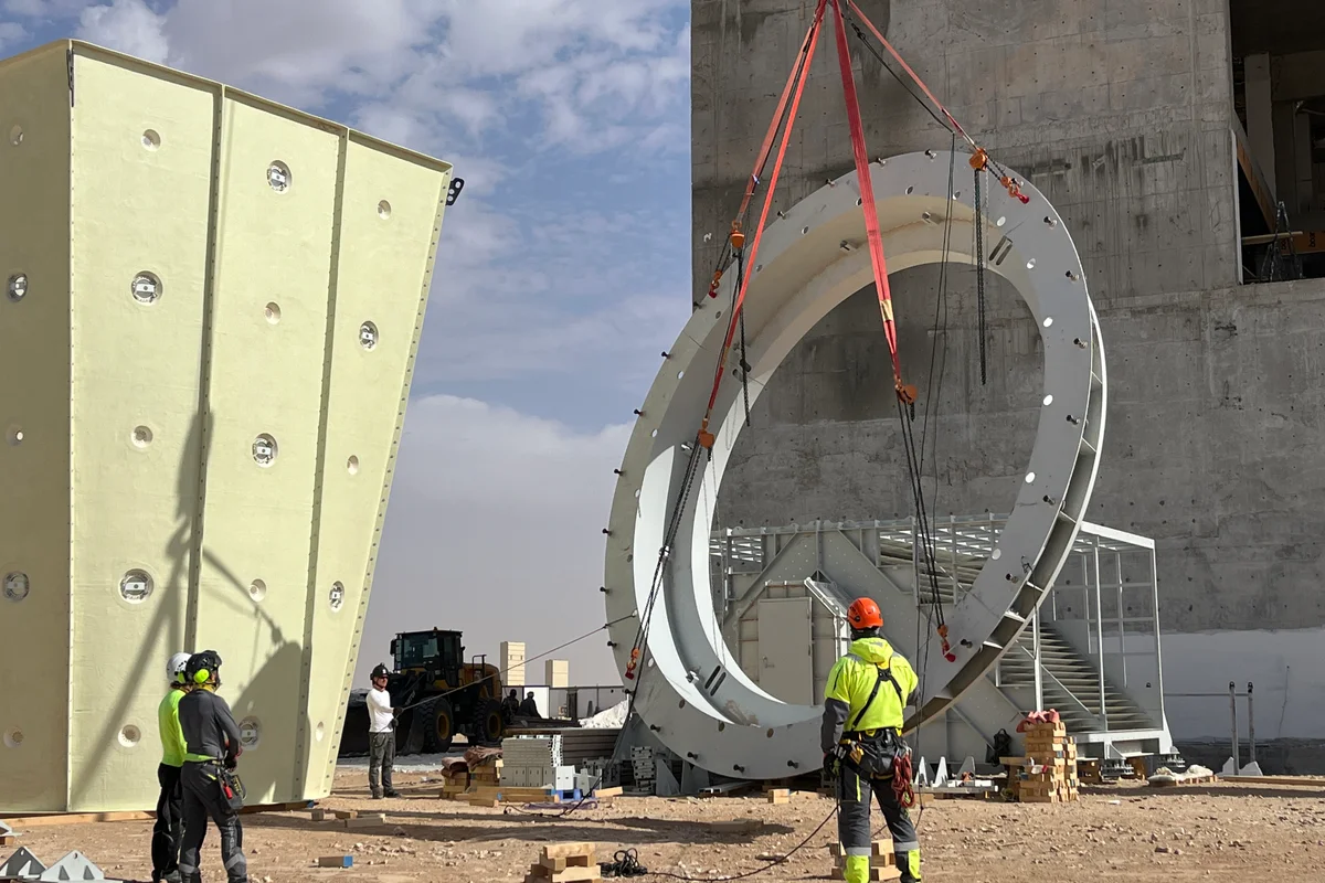 O pessoal de instalação guia o içamento por guindaste de um grande flange de aço estrutural no local de construção de um túnel de vento. Uma seção vertical do duto compósito de FRP da TunnelTech está à esquerda, apresentando a estrutura sanduíche projetada para absorção de ruído e eficiência de fluxo. O processo de montagem utiliza equipamentos de içamento pesado para posicionar componentes de grande diâmetro do circuito aerodinâmico.