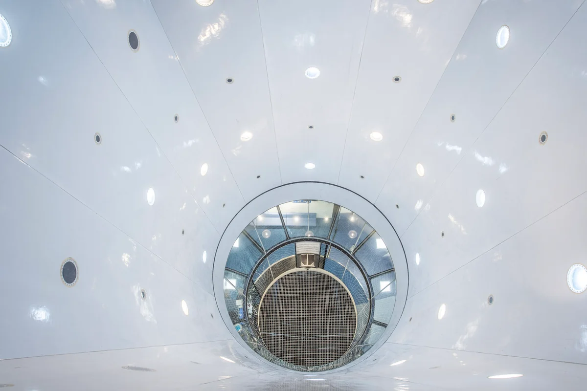 Een verticaal bovenaanzicht toont het interieur van de TT45 PRO windtunnel bij FlyStation Japan. Het perspectief kijkt door de verlengde witte diffusersectie, voorzien van inbouwverlichting, naar beneden in de 4.5-meter cilindrische glazen vliegruimte. Onderaan overspant het staalkabel-veiligheidsnet de luchtinlaat. Deze installatie beschikt over een verlengd bovenkamerontwerp, wat extra verticale ruimte biedt boven de primaire glassectie voor manoeuvres op grote hoogte.
