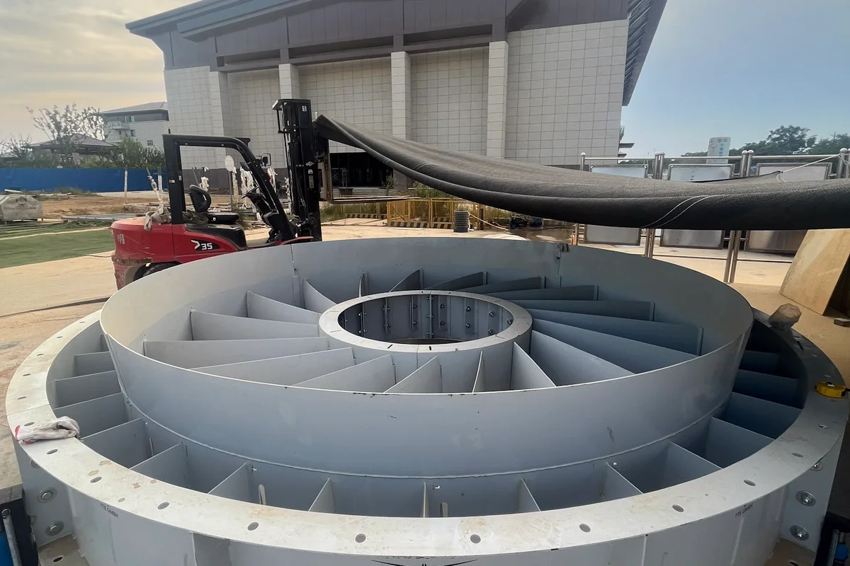 Sebuah rakitan baling-baling stator melingkar yang besar terletak di tanah selama instalasi terowongan angin TT43 Smart di China Flight Town. Komponen ini memiliki sirip pengarah radial tetap yang disusun di antara hub bagian dalam dan casing luar, dirancang untuk mengatur aliran udara di dalam bagian kipas aksial. Logo TTE GMBH terlihat di bagian luar rumah baja. Peralatan konstruksi, termasuk forklift dan perkakas listrik, mengelilingi unit tersebut dengan bangunan fasilitas sebagai latar belakang.
