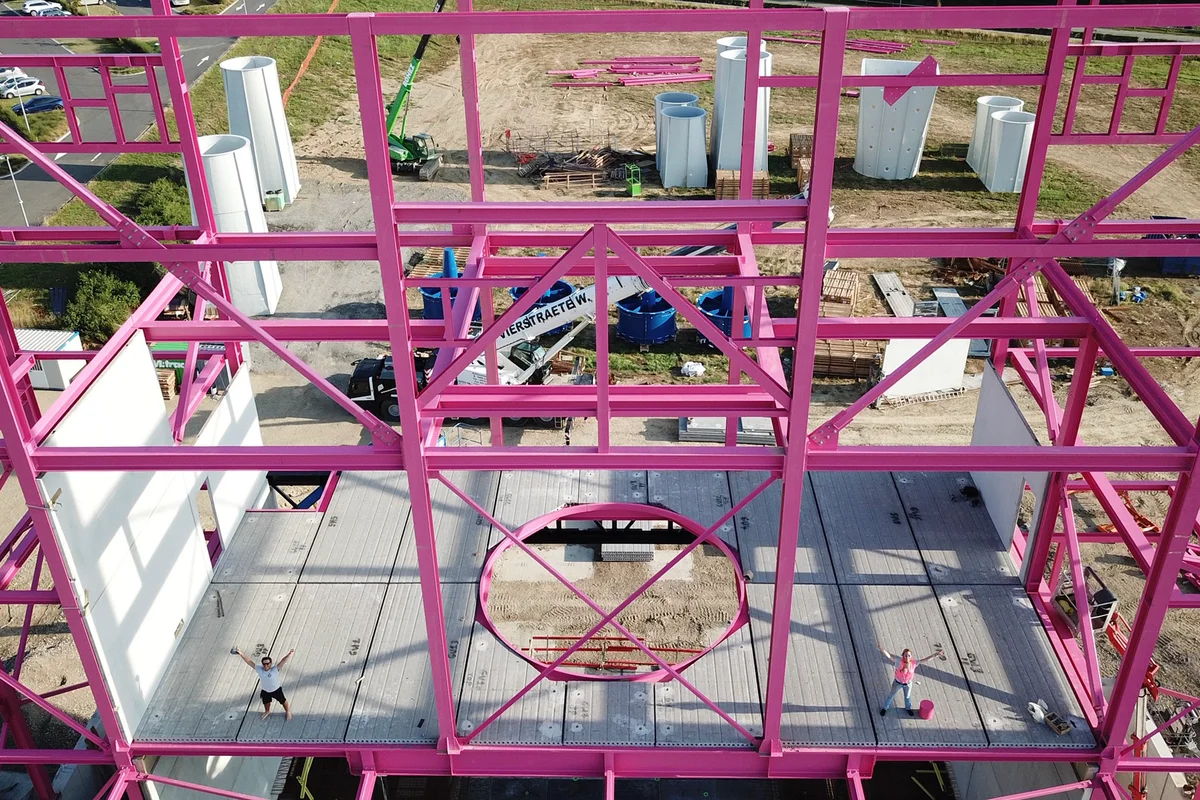 A imagem mostra o processo de montagem dos componentes estruturais do edifício Luxfly, incluindo as icónicas vigas de carga cor-de-rosa.