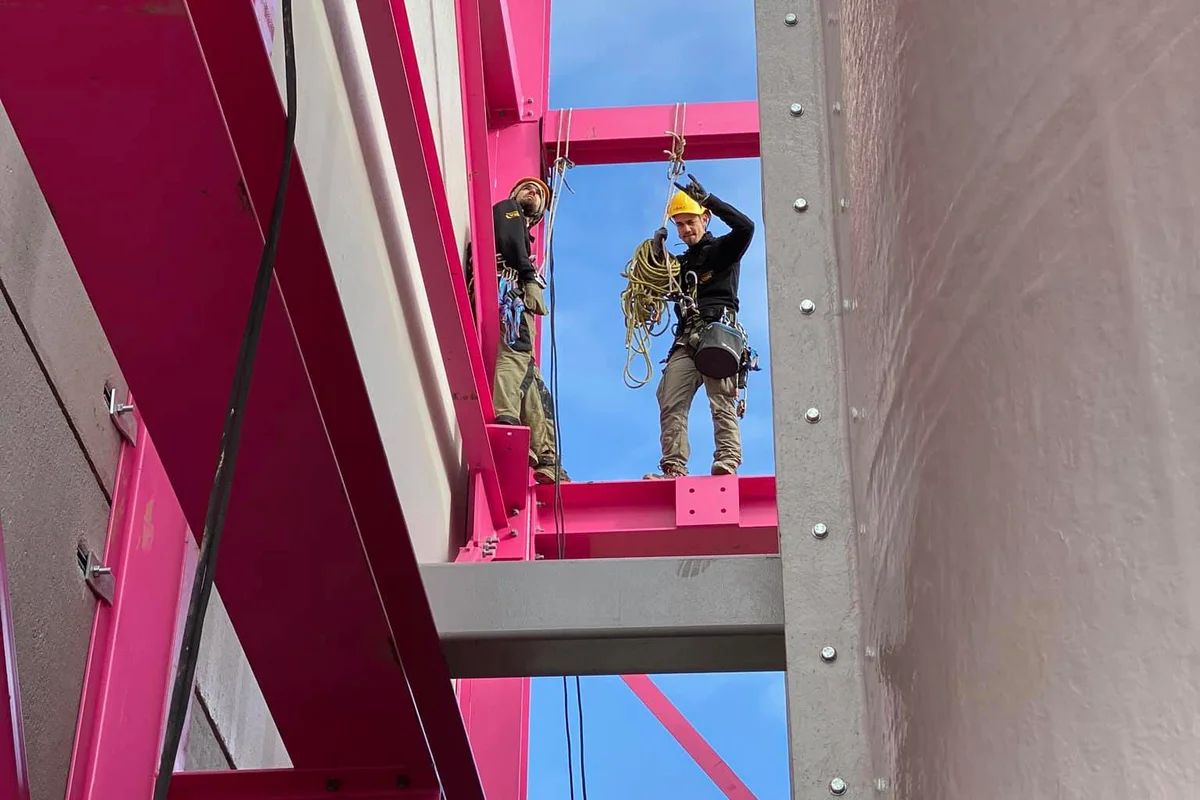 Hai kỹ thuật viên lắp đặt được trang bị dây đai an toàn và thiết bị buộc dây đứng trên khung thép kết cấu trong quá trình xây dựng cơ sở nhảy dù trong nhà Luxfly tại Luxembourg. Các nhân viên thực hiện các hoạt động buộc dây trên cao để căn chỉnh các thành phần hầm gió TunnelTech TT45 PRO với cấu trúc chịu lực màu hồng đặc trưng của tòa nhà. Ở bên phải, bề mặt bên ngoài của cụm hầm gió thẳng đứng có thể nhìn thấy liền kề với các dầm thép.