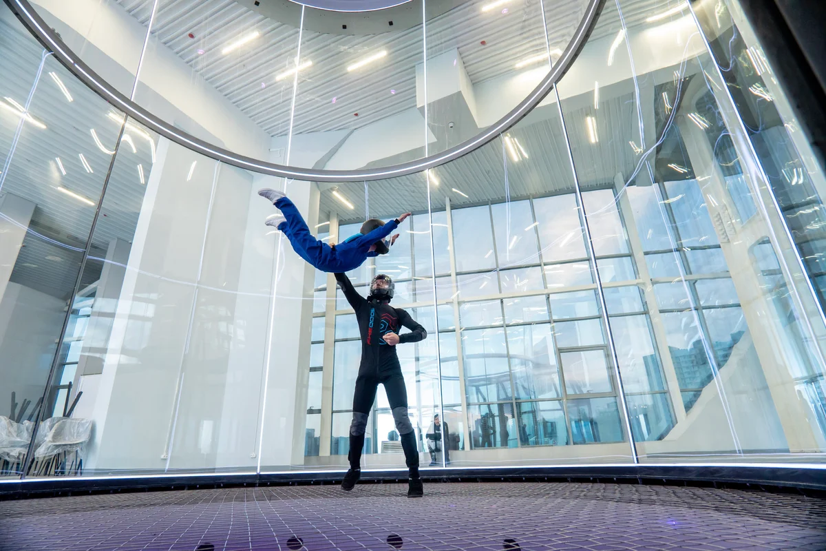 A professional instructor guides a young student through basic belly flying techniques inside the TT52 Pro at Kuzbas Arena. This 5.2m flight chamber features TunnelTech's signature frameless multilayer glass, offering spectators an unobstructed view of the action. You can see the effectiveness of the low-drag safety net as the instructor maintains precise control over the student's stability in the airflow.