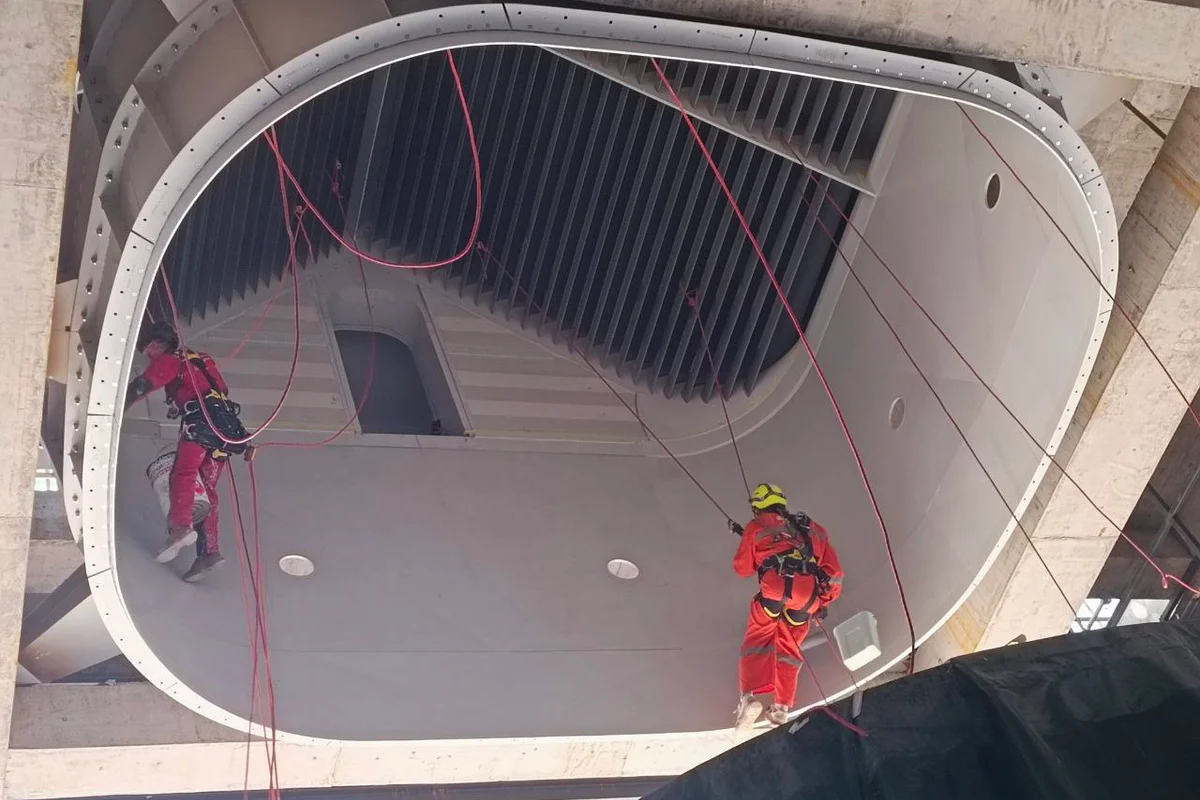 Teknikere udfører rope access-installation i den øvre sektion af flyvekammeret til TT43 Smart-vindtunnelen ved China Flight Town. Billedet viser den hvide kompositstruktur med cirkulære udskæringer designet til installation af lyskilder. Over arbejderne giver en rektangulær dør adgang til BASE jumping-kammeret. Øverst i sektionen er ledeskovle arrangeret i en V-formet konfiguration for at dele luftstrømmen i venstre og højre returkanaler.