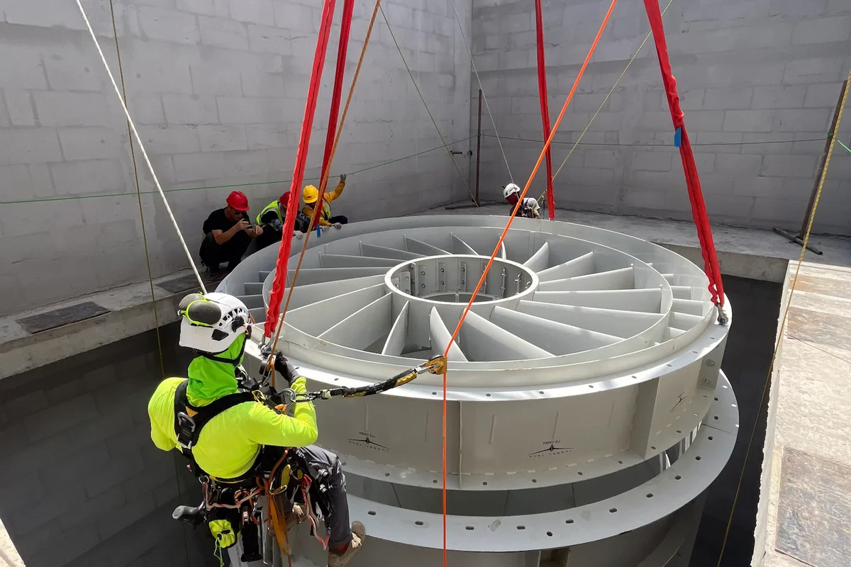 Aukštalipis technikas vadovauja ašinio ventiliatoriaus statoriaus menčių bloko montavimui į betoninį korpusą. Ši sunkiasvorė kėlimo operacija yra TunnelTech TT43 Smart vėjo tunelio statybos dalis China Flight Town projekte. Plieninėje sekcijoje yra stacionarios statoriaus mentės, reikalingos oro srautui ištiesinti vertikaliame grįžtamajame ortakyje. Komponentas kabo ant raudonų kėlimo diržų, leidžiantis į objekto betoninę konstrukciją.