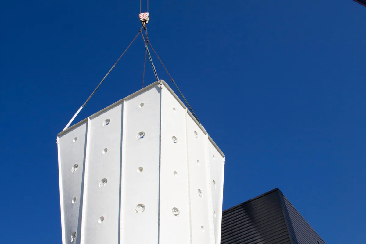 Een mobiele kraan hijst een wit, gefacetteerd FRP (glasvezel) diffusersegment op zijn plaats bij de faciliteit Windalps in Frankrijk. Dit onderdeel vormt de bovenste sectie van de vluchtkamer, direct boven de glazen vliegzone. De structuur bevat duidelijke cirkelvormige poorten die zijn ontworpen voor de installatie van verlichtingselementen. Als onderdeel van het recirculatiecircuit vergroot deze diffuser de dwarsdoorsnede van de luchtstroom om de luchtsnelheid te verlagen wanneer deze de vluchtkamer verlaat.