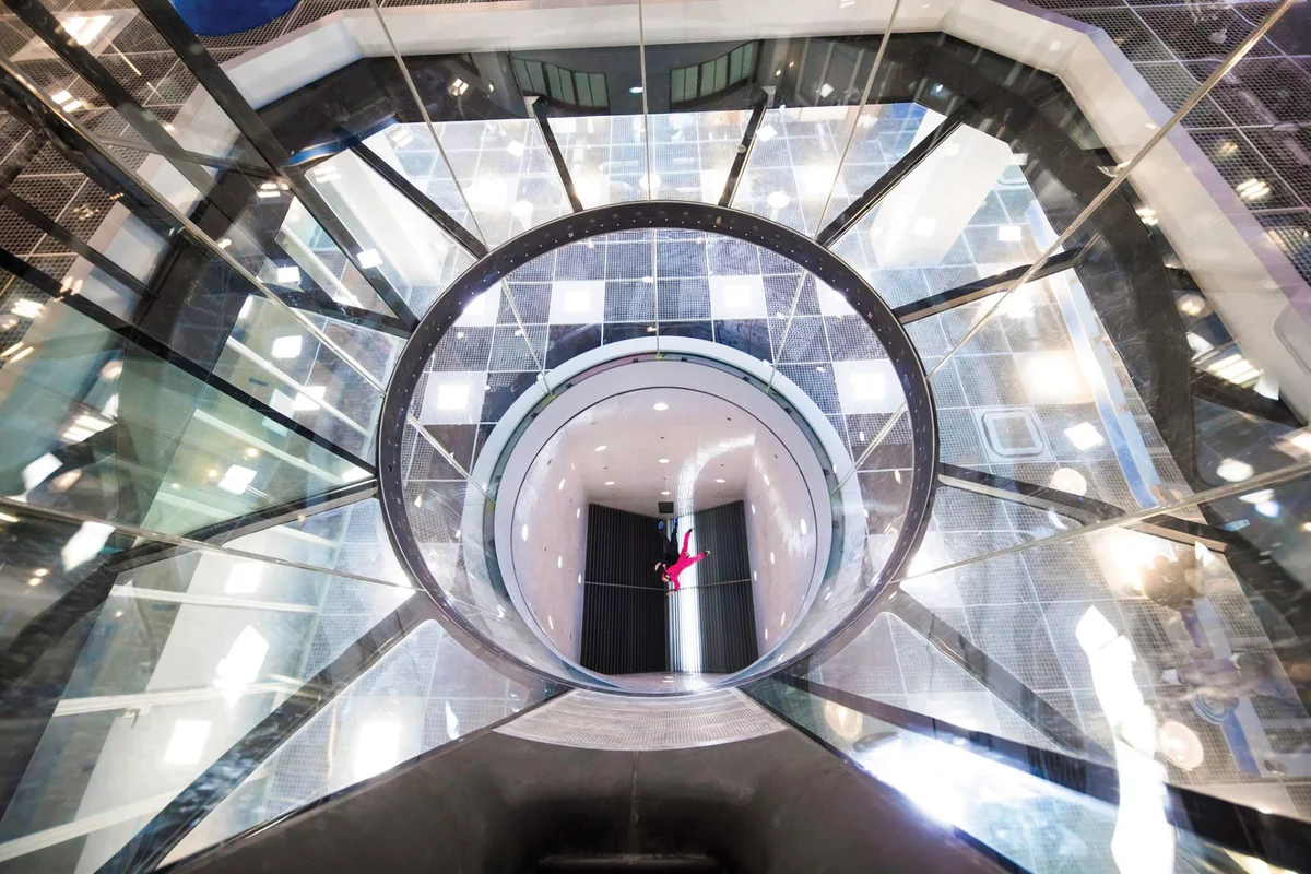 This low-angle view captures the flight chamber from the safety net looking upward. An instructor is performing a taxi flight with a student near the top of the chamber, close to the turning vanes and the base jumping camera. The pair is flying at a height of approximately 20 meters. The perspective highlights the vertical scale of the glass walls and the architectural framework surrounding the airflow column.