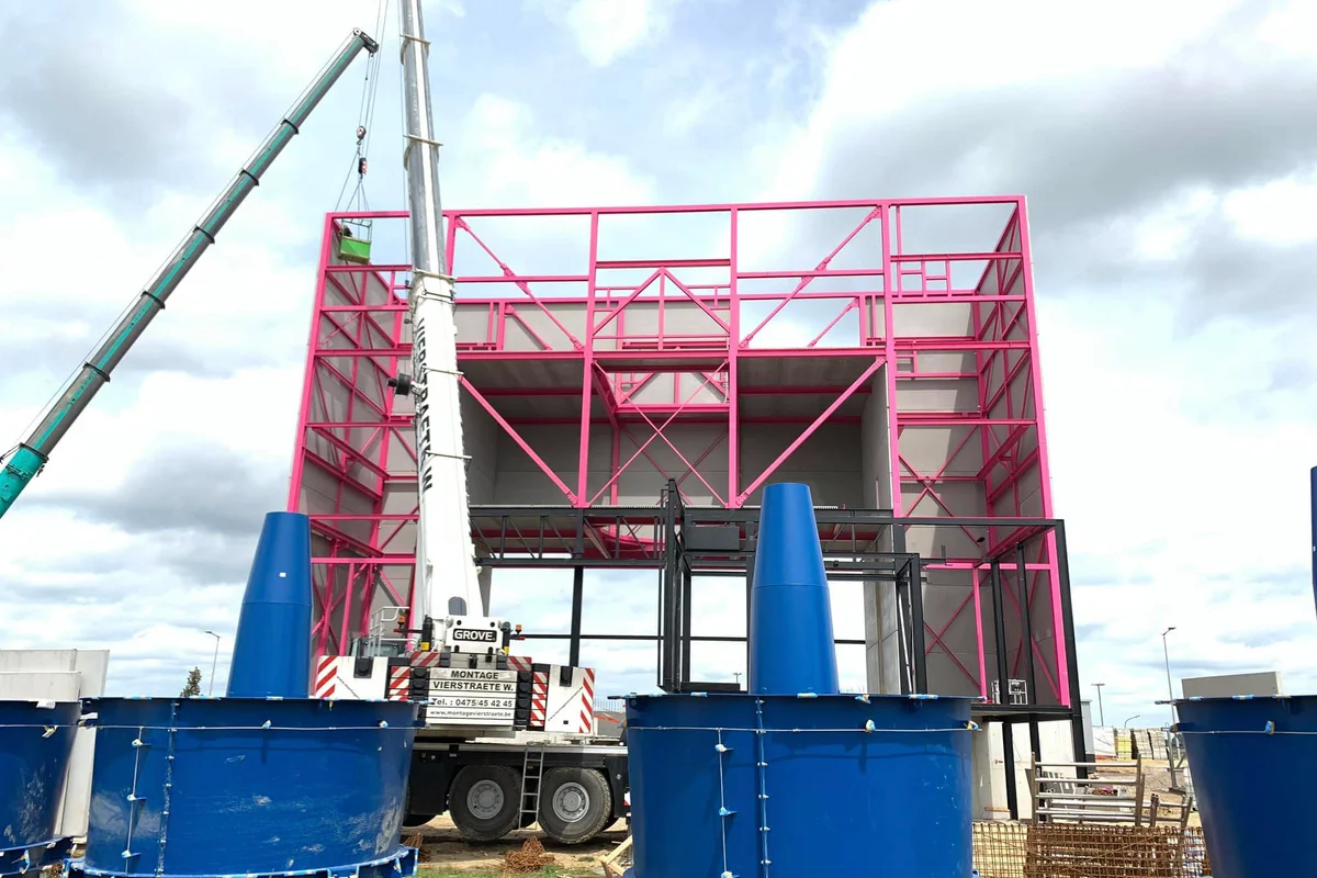 Ang malalaking asul na fan housing units na naglalaman ng stator vanes ay naghihintay ng installation sa site ng Luxfly wind tunnel sa Luxembourg. Isang mobile crane ang gumagana katabi ng natatanging pink na steel bearing structure ng pasilidad. Ang mabibigat na mekanikal na bahaging ito ay bumubuo sa static na seksyon ng axial fan assembly para sa sistemang TT45 PRO. Inihahanda ng construction team ang kagamitan sa lupa bago ito iangat sa return duct loop ng pasilidad.